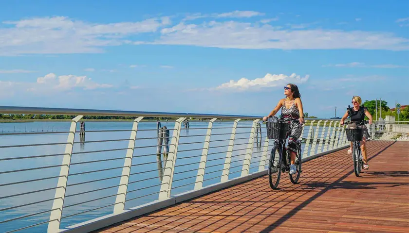 Due donne in bicicletta su una moderna pista ciclabile jesi, con il mare e il cielo azzurro sullo sfondo.
