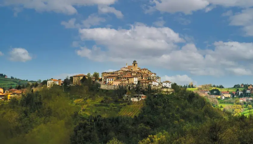 Borgo antico su un colle, con case in pietra e tetti rossi, sotto un cielo azzurro con nuvole bianche. Il **monte Grimano** domina il paesaggio.
