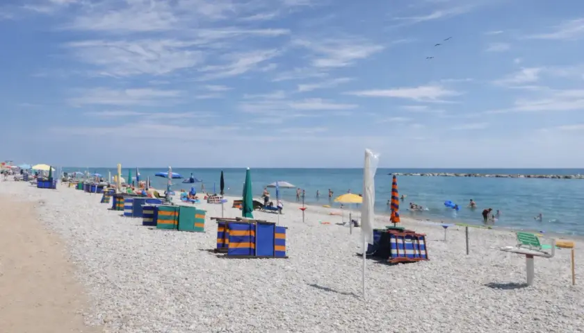 Spiaggia di ciottoli con ombrelloni colorati e cabine, bagnanti in mare. Un tranquillo fermo mare sotto un cielo azzurro con nuvole leggere.