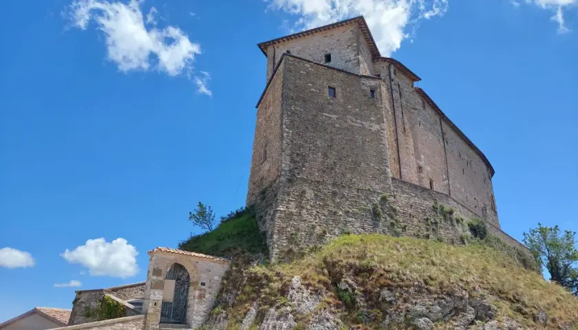 Antico borgo di Frontone, Marche, con imponente rocca in pietra su un colle erboso, sotto un cielo azzurro con nuvole bianche.