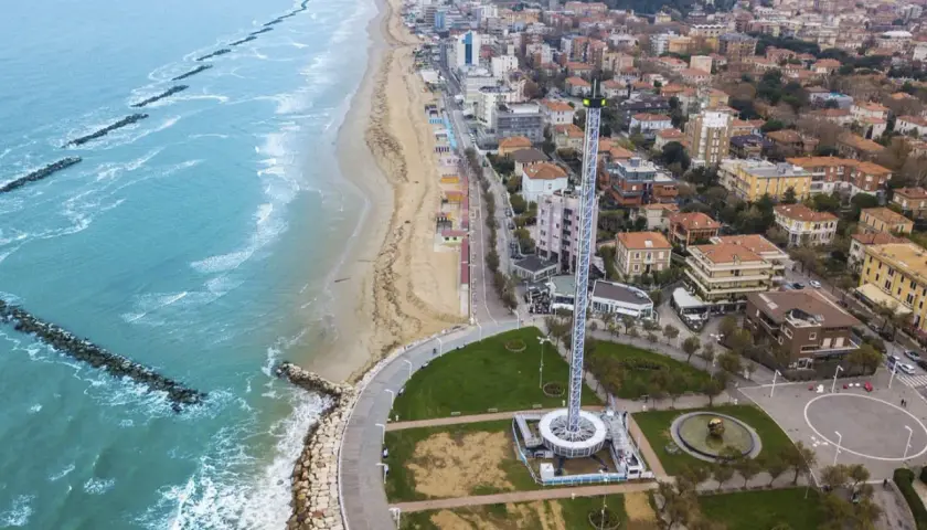 Vista aerea del lungomare di Pesaro con la spiaggia, il mare con frangiflutti e una torre panoramica.