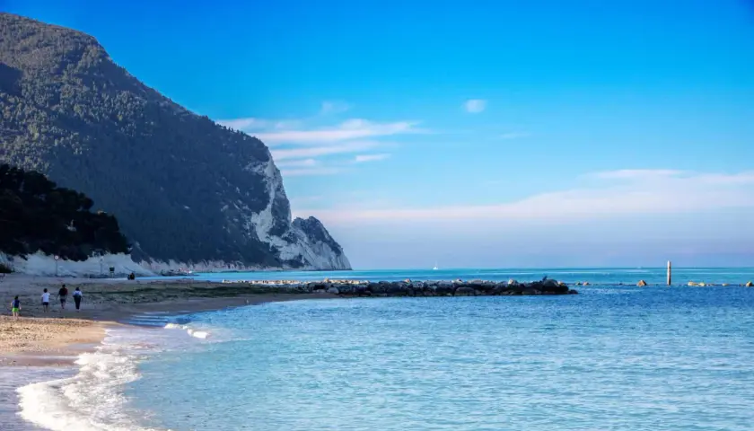Scogliera a picco sul mare azzurro, spiaggia dorata e gente che passeggia sulla riviera marchigiana.