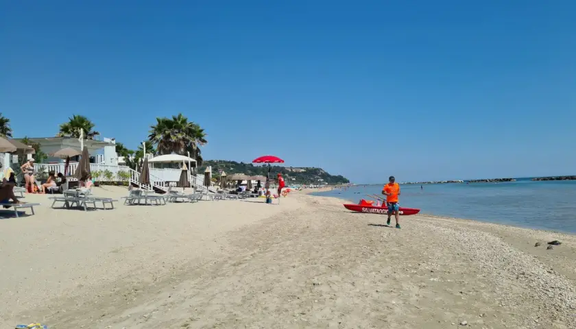 Sole splendente sulla campofilone spiaggia, con bagnanti rilassati, ombrelloni e un bagnino pronto a intervenire vicino a una barca di salvataggio.