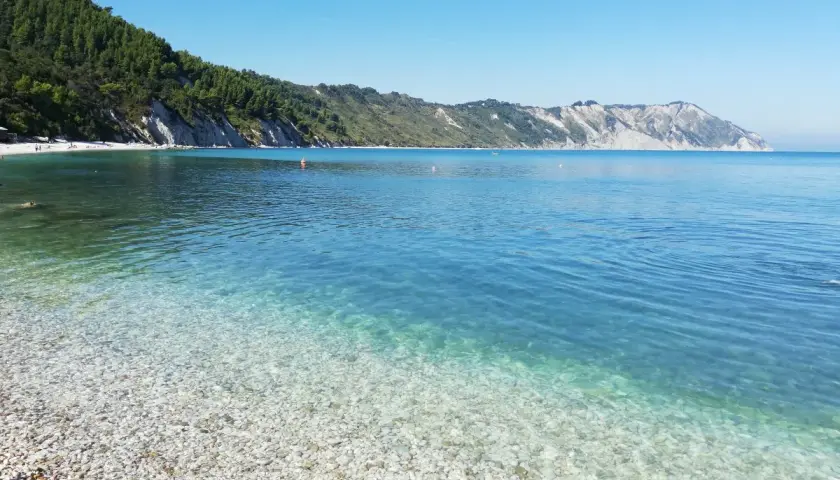 Acque cristalline e ciottoli bianchi a Portonovo, una delle spiagge più belle d'Italia. La costa verdeggiante si tuffa nel mare azzurro.