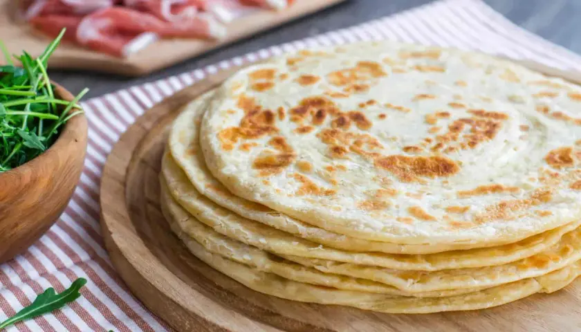 Una pila di cresce dorate su un tagliere di legno, pronte per essere farcite con rucola fresca e prosciutto.