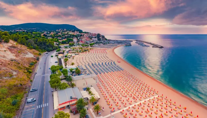 Vista aerea della spiaggia numana con file ordinate di ombrelloni rossi e blu, mare calmo e cielo rosa al tramonto.