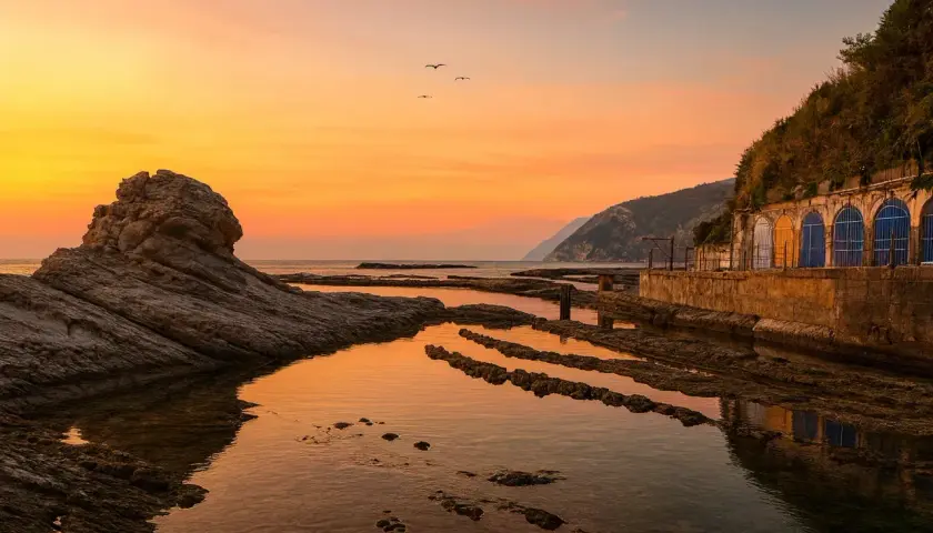 Tramonto dorato sulla spiaggia del Passetto, con rocce scolpite e archi colorati che si riflettono nell'acqua calma.