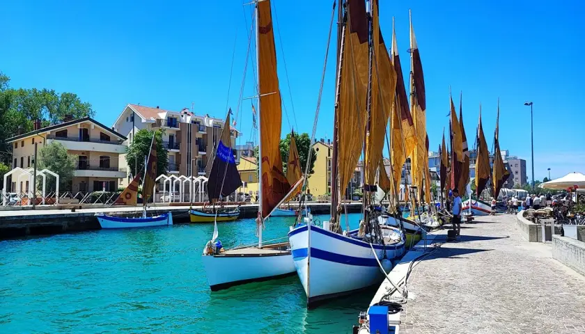 Barche a vela ormeggiate in un porto con case colorate sullo sfondo. Un dilemma: meglio Bellaria o Igea Marina?