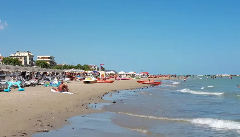Spiaggia affollata a Cupra Marittima, con ombrelloni, lettini, bagnanti e barche di salvataggio. Il mare azzurro invita a un tuffo.