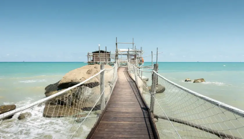 Ponte in legno verso un trabucco, tipica struttura da pesca delle Marche, su un mare azzurro e limpido.