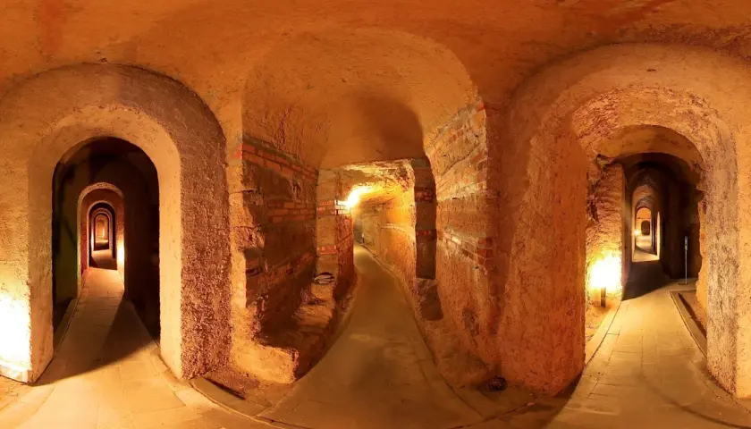 Un labirinto di corridoi illuminati, con archi in mattoni e terra battuta, che si estende in profondità. Sembra di esplorare antiche grotte sotterranee.