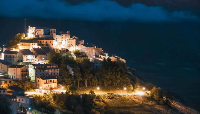 Norcia: cosa vedere dopo il terremoto. Borgo illuminato di notte, arroccato su un colle, con case in pietra e alberi.