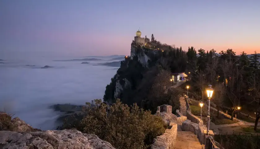 Torre Guaita domina la nebbia al tramonto, un invito a visitare San Marino per un'esperienza magica.