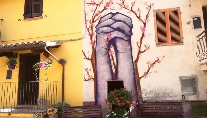 Braccano, paese dei murales: mani giunte e fiori di ciliegio dipinti su un muro, con panchine e decorazioni in legno.