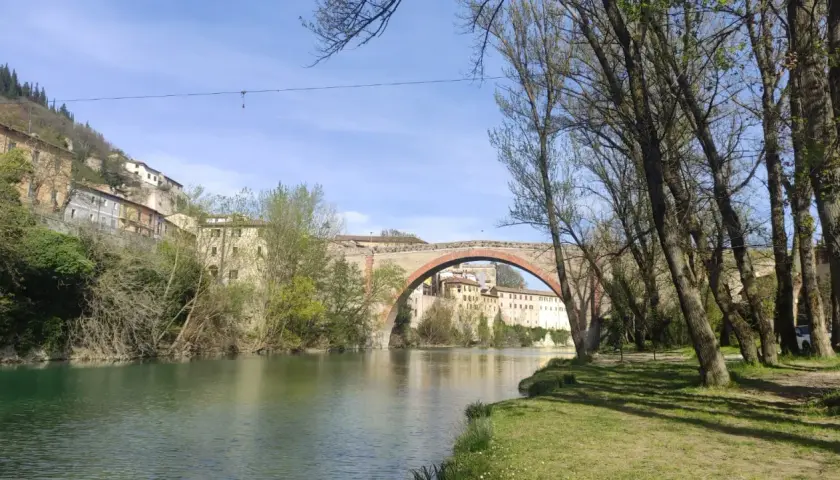 Il fiume scorre placido sotto il ponte ad arco del canyon di Fossombrone, con case antiche aggrappate alla collina e alberi che incorniciano la scena.