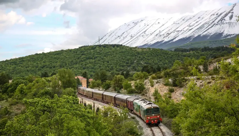 Treno storico attraversa un ponte in pietra, circondato da verde lussureggiante e montagne innevate. Un viaggio suggestivo sulla Transiberiana d'Italia.