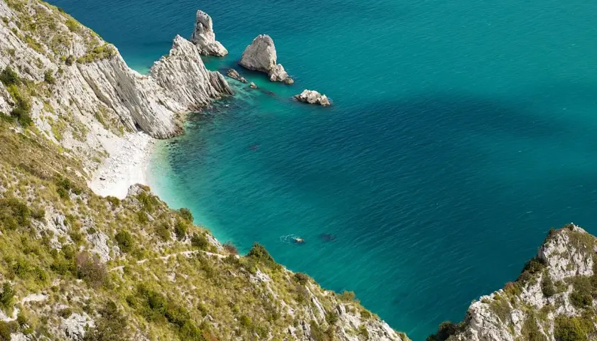 Scogliere bianche e mare cristallino del Conero, con una piccola spiaggia nascosta e un nuotatore solitario.