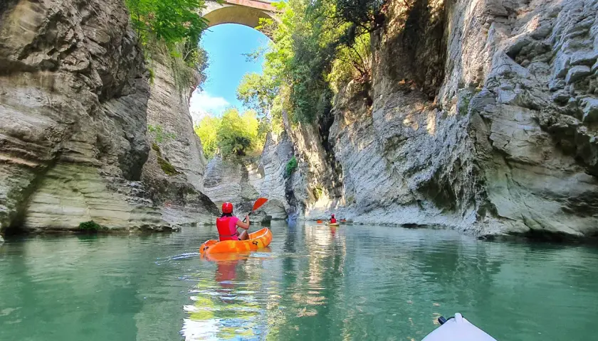 Persone in canoa navigano un fiume tra alte pareti rocciose, un paesaggio che ricorda le marmitte dei giganti, sotto un antico ponte ad arco.