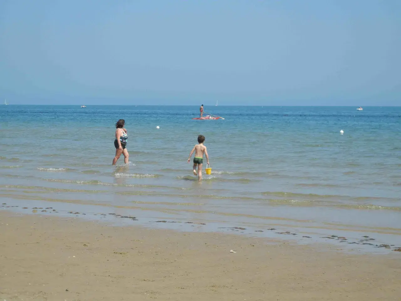 Bambino con secchiello giallo corre verso il mare a Pesaro, mentre una donna cammina nell'acqua. In lontananza, un uomo su una tavola da SUP.