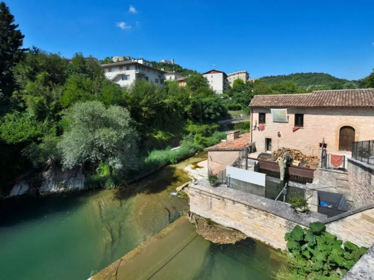 Sassoferrato cosa vedere: un antico mulino ad acqua lungo un fiume, con case e vegetazione lussureggiante sullo sfondo.