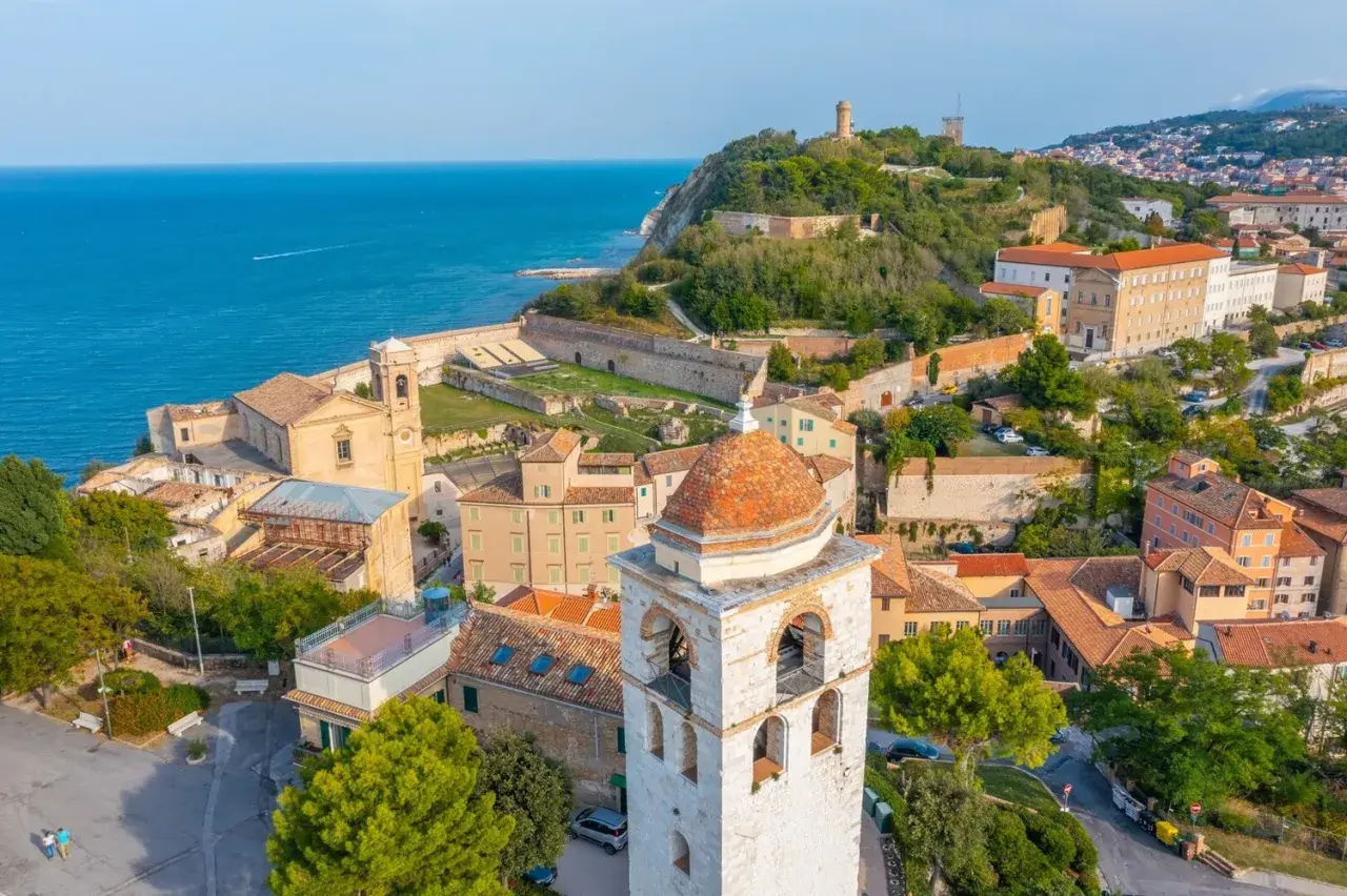 Vista aerea del **centro storico** di Ancona, con il mare blu, chiese, torri e case con tetti rossi.