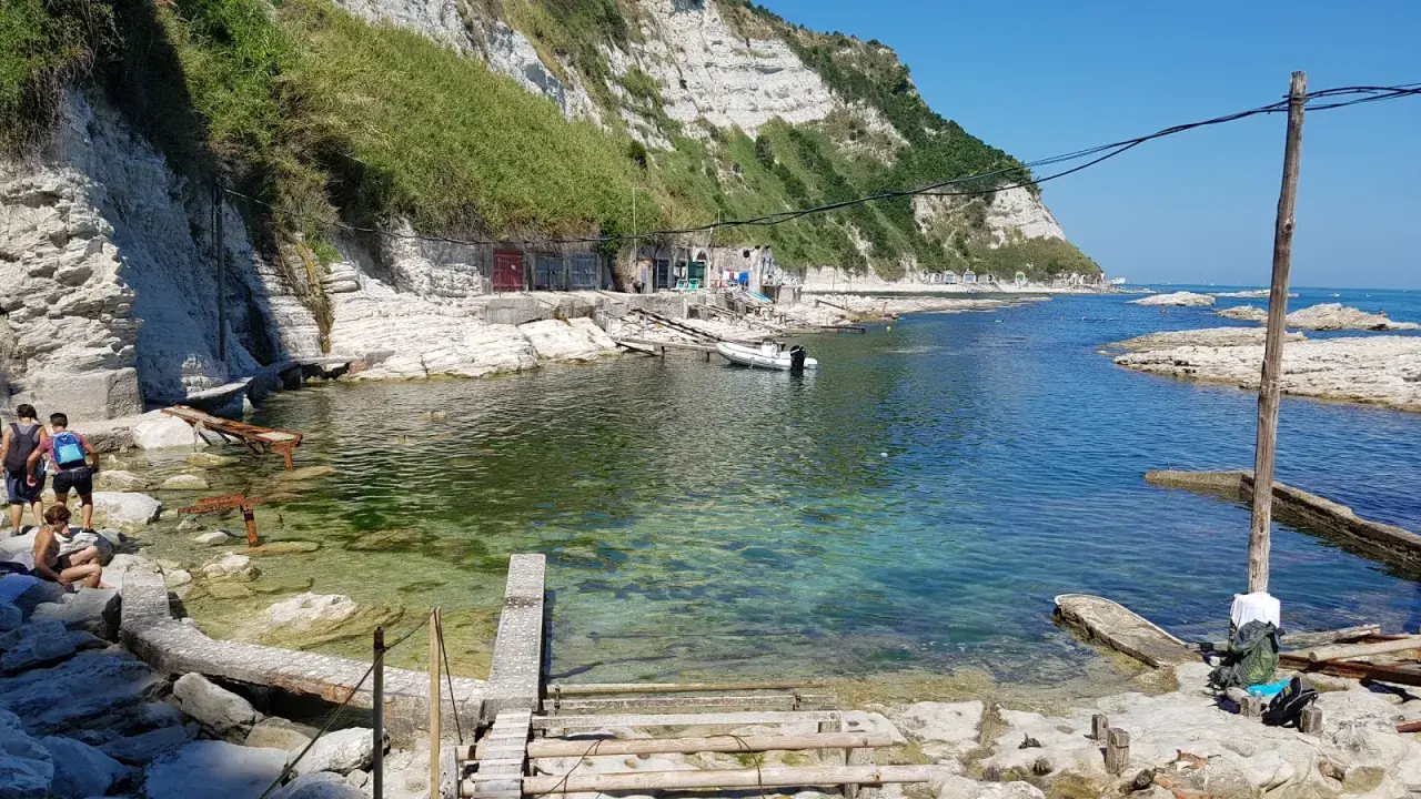 Scogli bianchi e mare cristallino alla spiaggia della Scalaccia. Una piccola barca è ormeggiata vicino a casotti colorati.