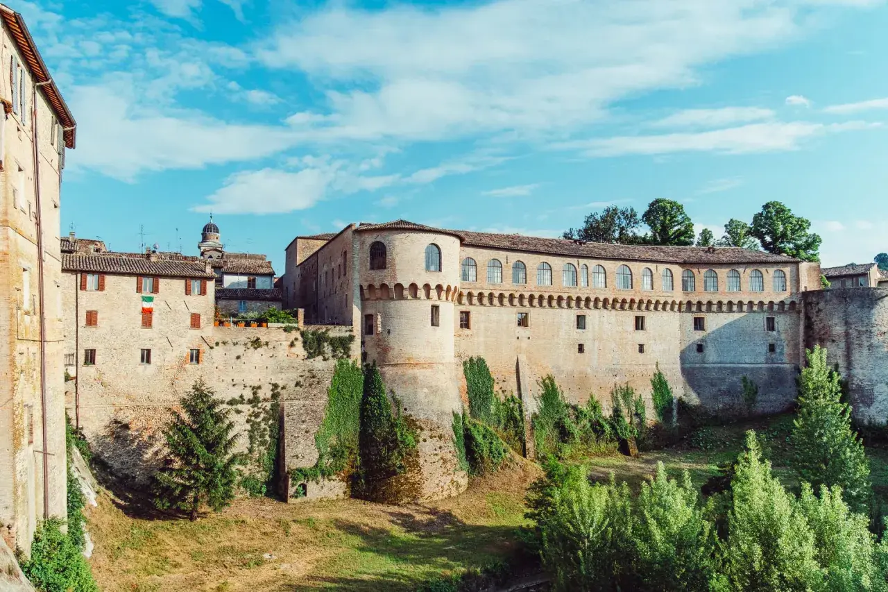 Un imponente palazzo storico con torri e mura di mattoni, circondato da vegetazione. Un luogo da scoprire: Urbania cosa vedere.