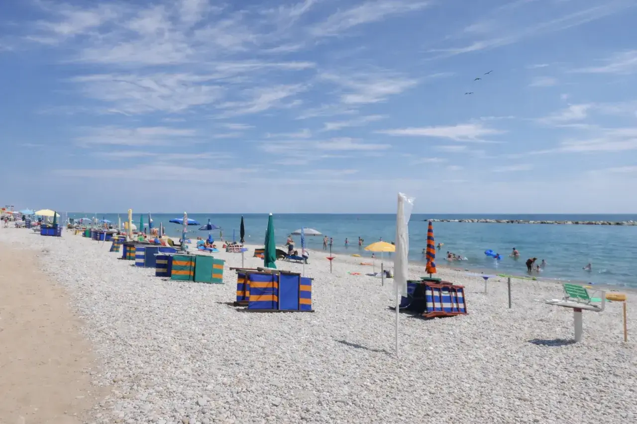 Spiaggia di ciottoli con ombrelloni colorati e cabine, bagnanti in mare. Un tranquillo fermo mare sotto un cielo azzurro con nuvole leggere.