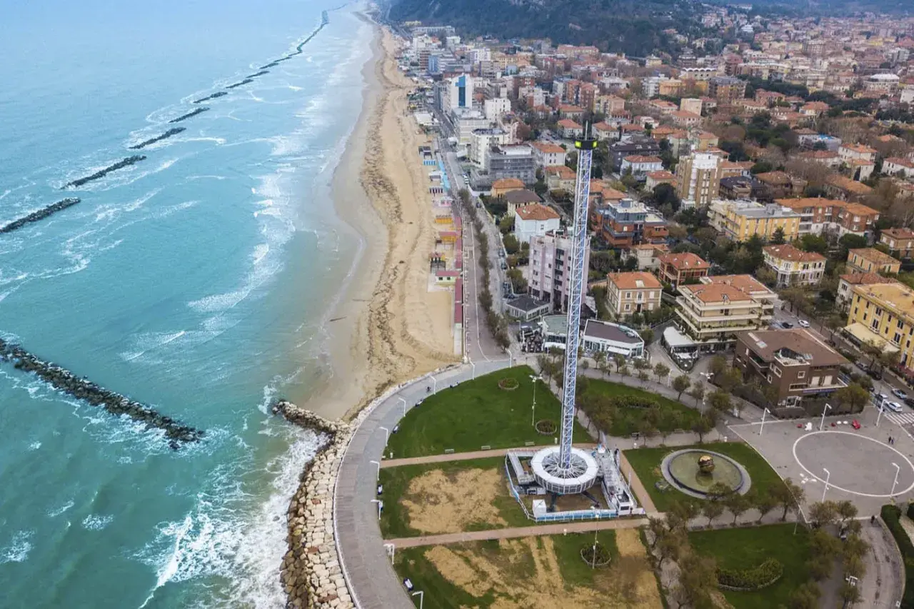 Vista aerea del lungomare di Pesaro con la spiaggia, il mare con frangiflutti e una torre panoramica.