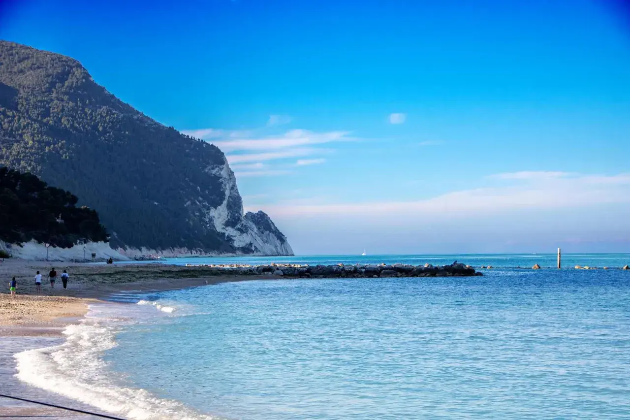 Scogliera a picco sul mare azzurro, spiaggia dorata e gente che passeggia sulla riviera marchigiana.