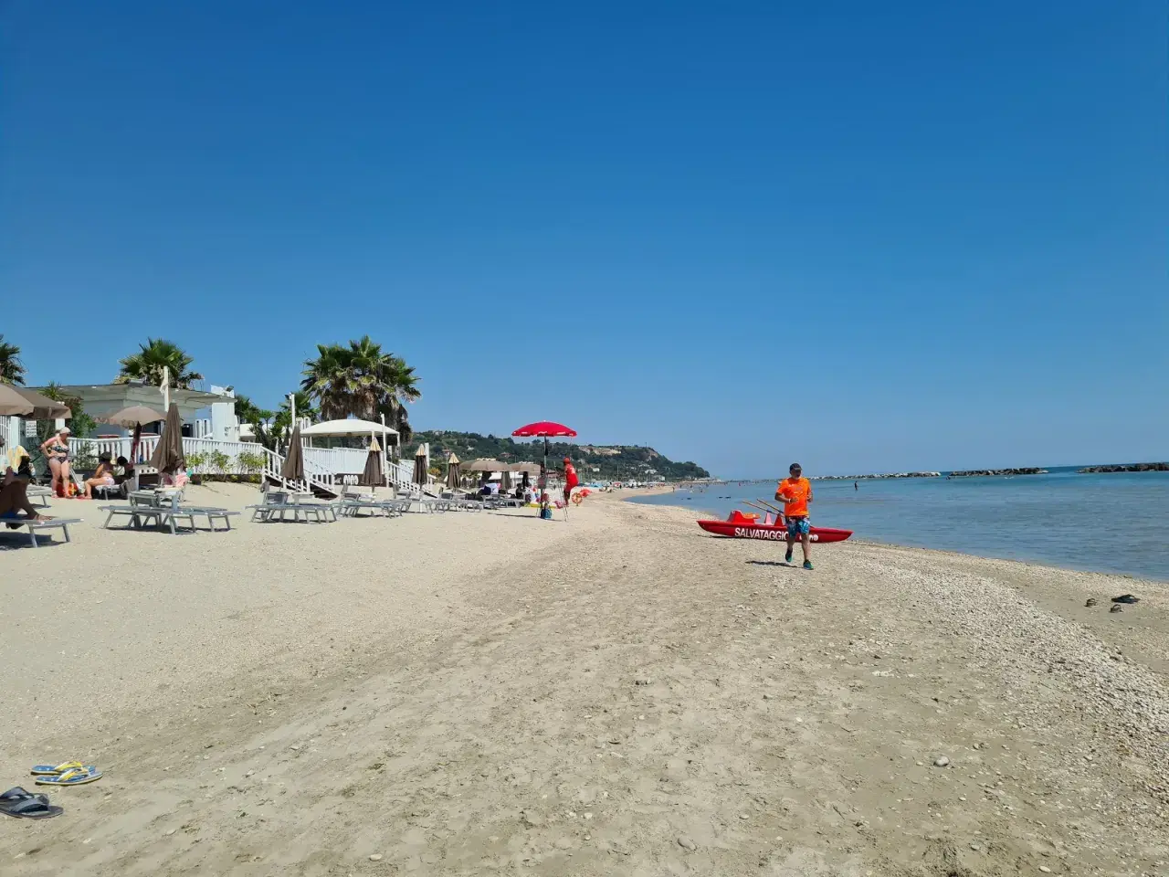 Sole splendente sulla campofilone spiaggia, con bagnanti rilassati, ombrelloni e un bagnino pronto a intervenire vicino a una barca di salvataggio.