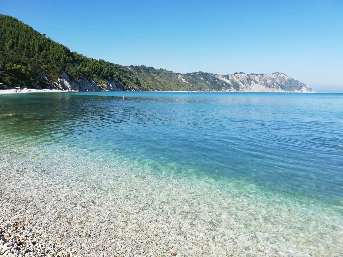 Acque cristalline e ciottoli bianchi a Portonovo, una delle spiagge più belle d'Italia. La costa verdeggiante si tuffa nel mare azzurro.