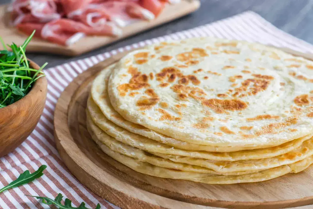 Una pila di cresce dorate su un tagliere di legno, pronte per essere farcite con rucola fresca e prosciutto.