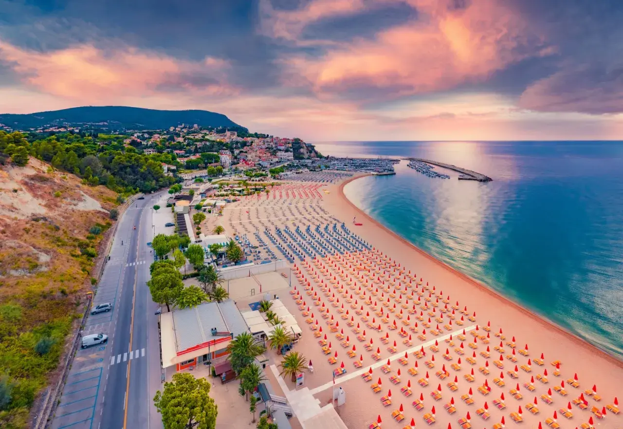 Vista aerea della spiaggia numana con file ordinate di ombrelloni rossi e blu, mare calmo e cielo rosa al tramonto.