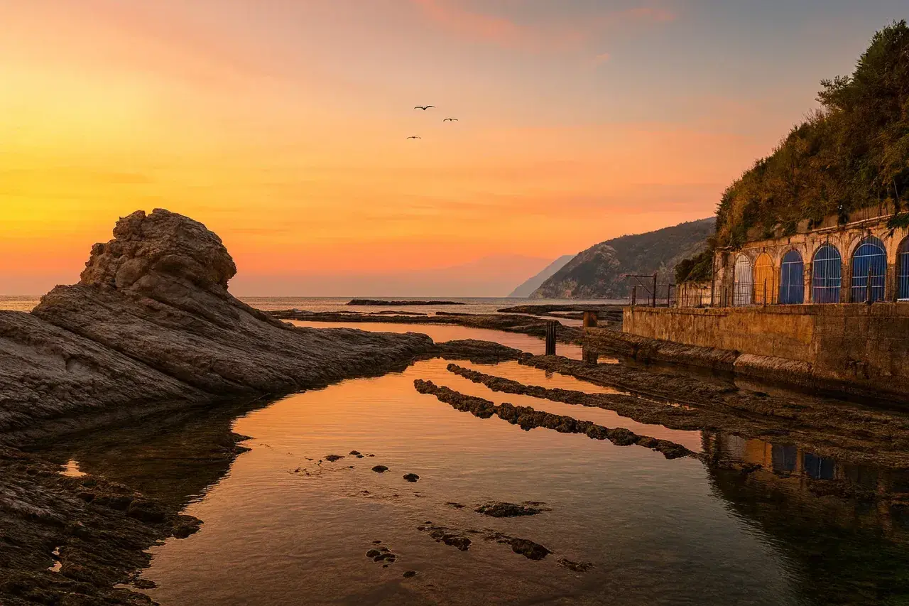 Tramonto dorato sulla spiaggia del Passetto, con rocce scolpite e archi colorati che si riflettono nell'acqua calma.