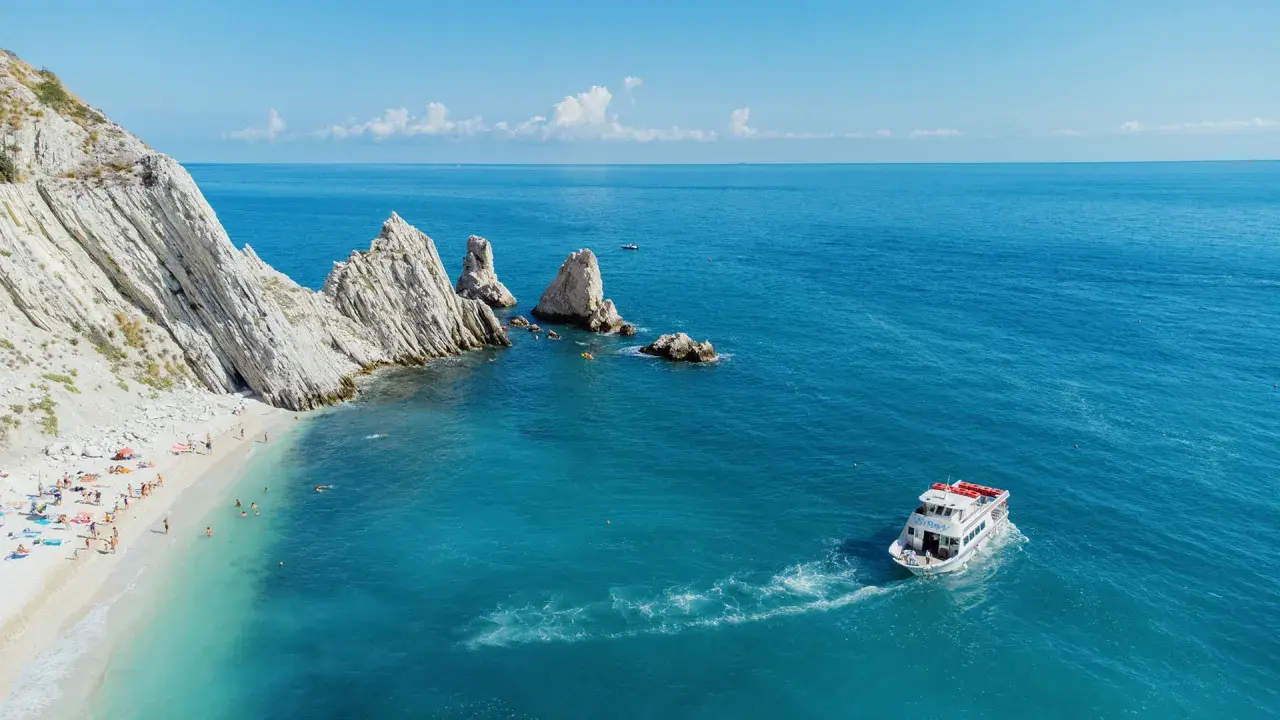 La spiaggia delle Due Sorelle, con le sue iconiche rocce, accoglie bagnanti e kayak. Una barca solca il mare azzurro.