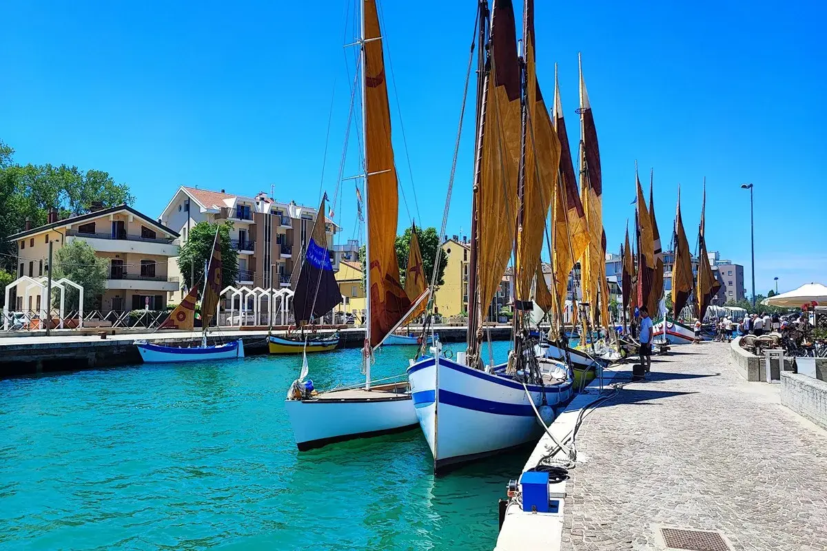 Barche a vela ormeggiate in un porto con case colorate sullo sfondo. Un dilemma: meglio Bellaria o Igea Marina?