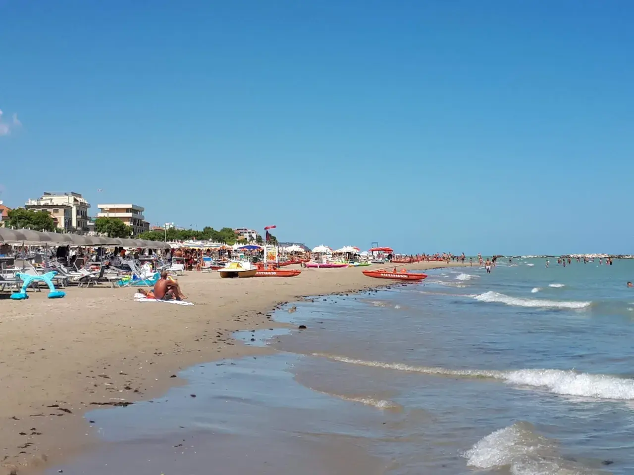 Spiaggia affollata a Cupra Marittima, con ombrelloni, lettini, bagnanti e barche di salvataggio. Il mare azzurro invita a un tuffo.