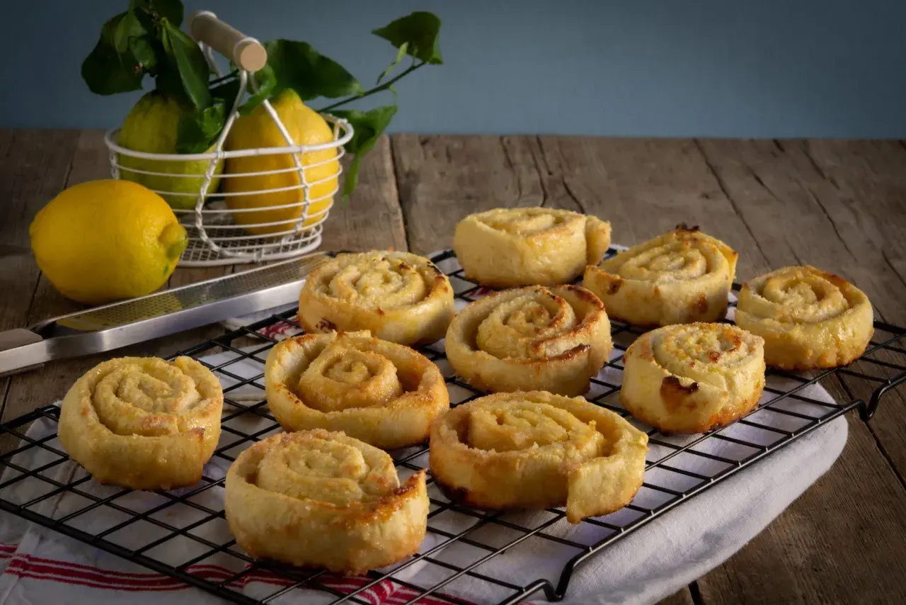 Dolci rotolini profumati ai limoni, perfetti per il Carnevale. Freschi limoni in un cestino e una grattugia completano la scena.
