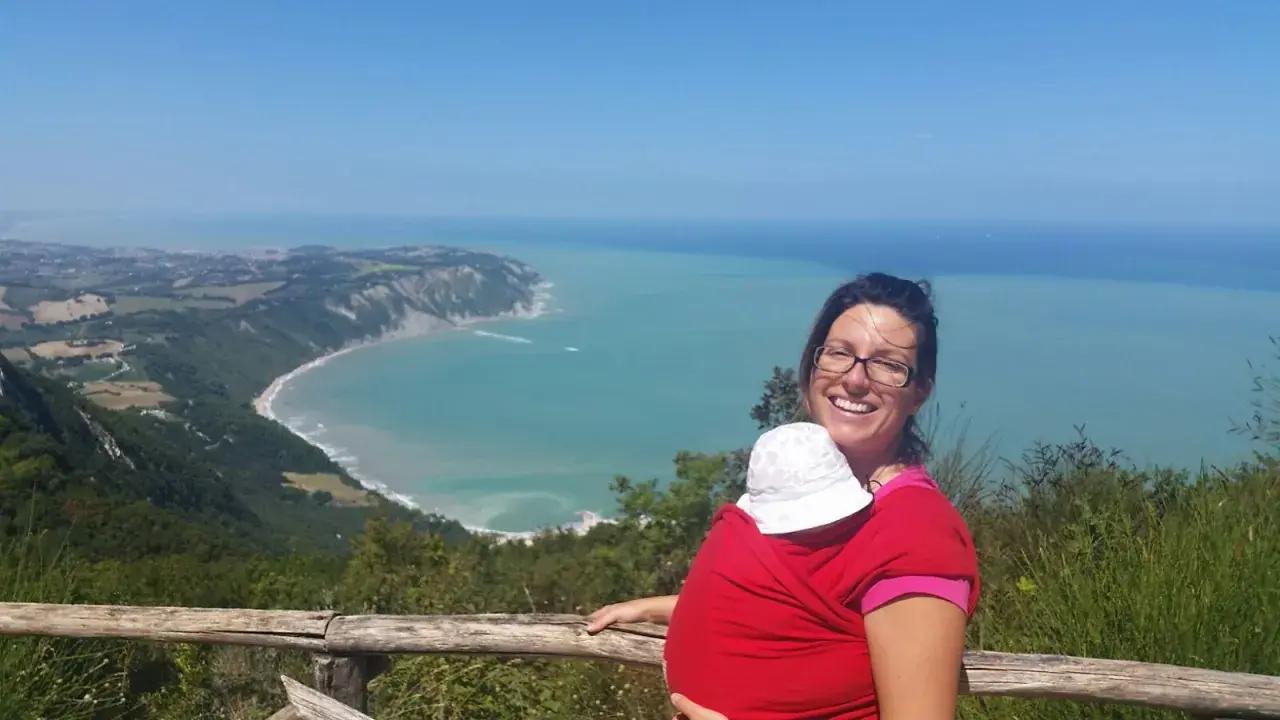 Donna sorridente con bimbo in fascia, ammira la costa del Conero dai sentieri. Perfetto per esplorare i sentieri del Conero con bambini.