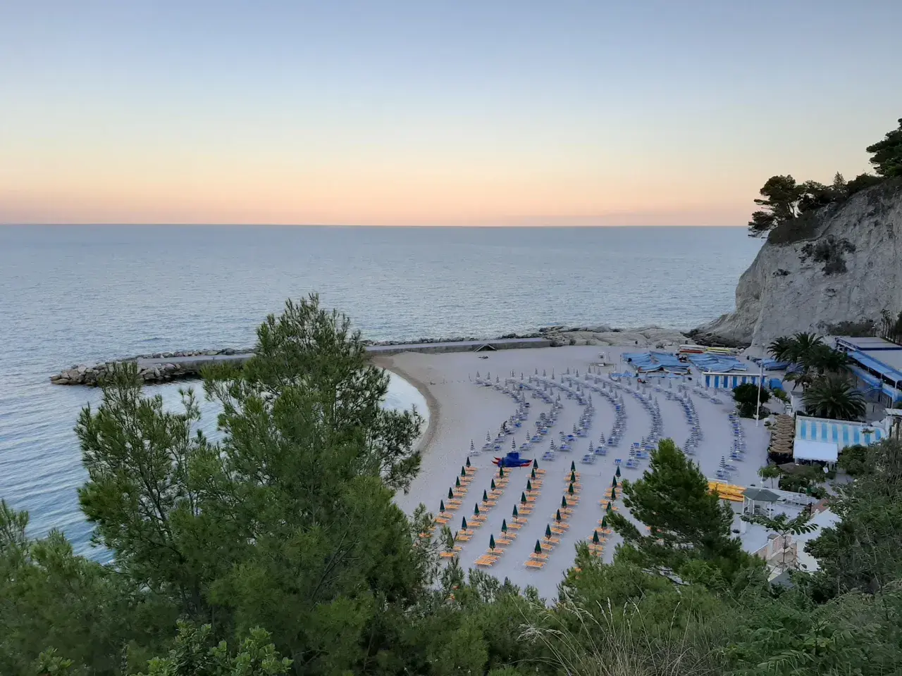 Spiaggia ordinata con lettini e ombrelloni, ideale per scoprire cosa vedere a Numana e Sirolo. Mare calmo al tramonto.
