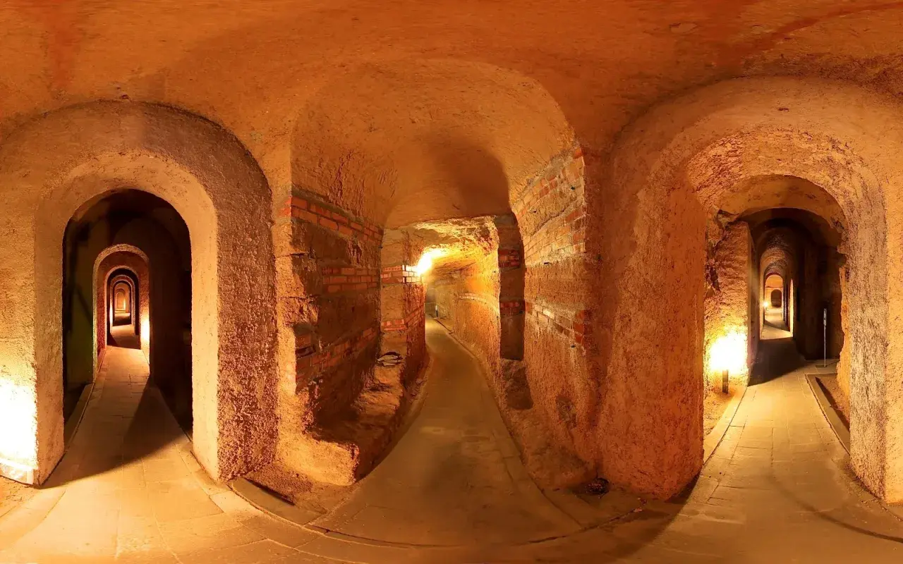 Un labirinto di corridoi illuminati, con archi in mattoni e terra battuta, che si estende in profondità. Sembra di esplorare antiche grotte sotterranee.