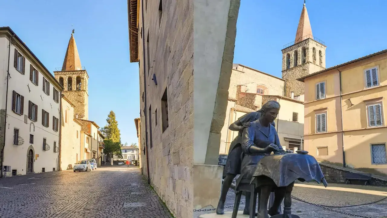 Sansepolcro cosa vedere: una piazza con una scultura di donne al lavoro, campanili medievali e palazzi storici sotto un cielo azzurro.