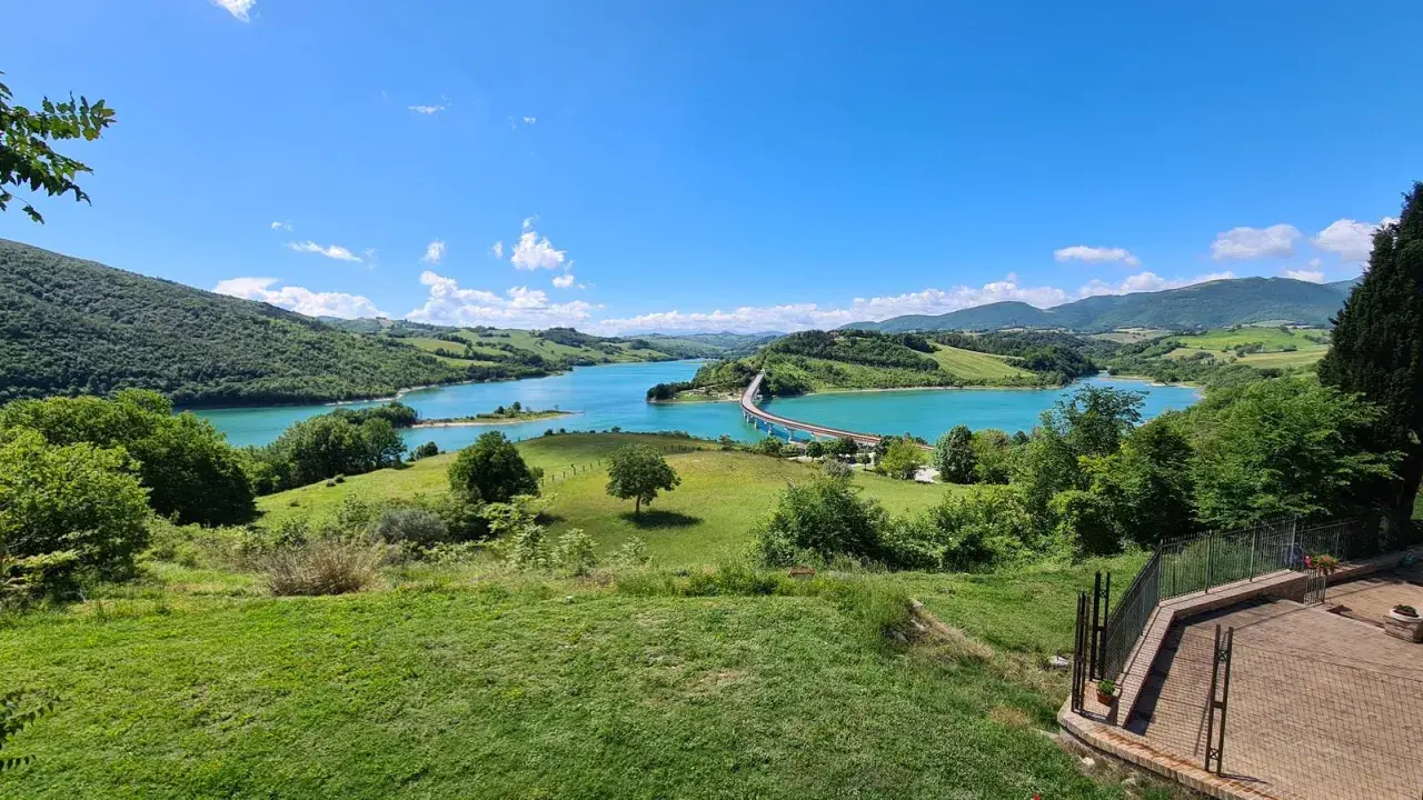 Panoramica della diga di Cingoli con lago turchese, colline verdi e un ponte ad arco sotto un cielo azzurro.