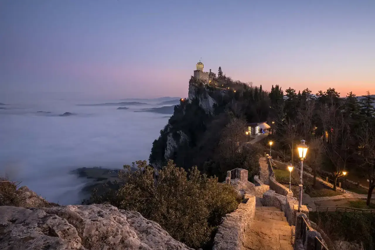 Torre Guaita domina la nebbia al tramonto, un invito a visitare San Marino per un'esperienza magica.