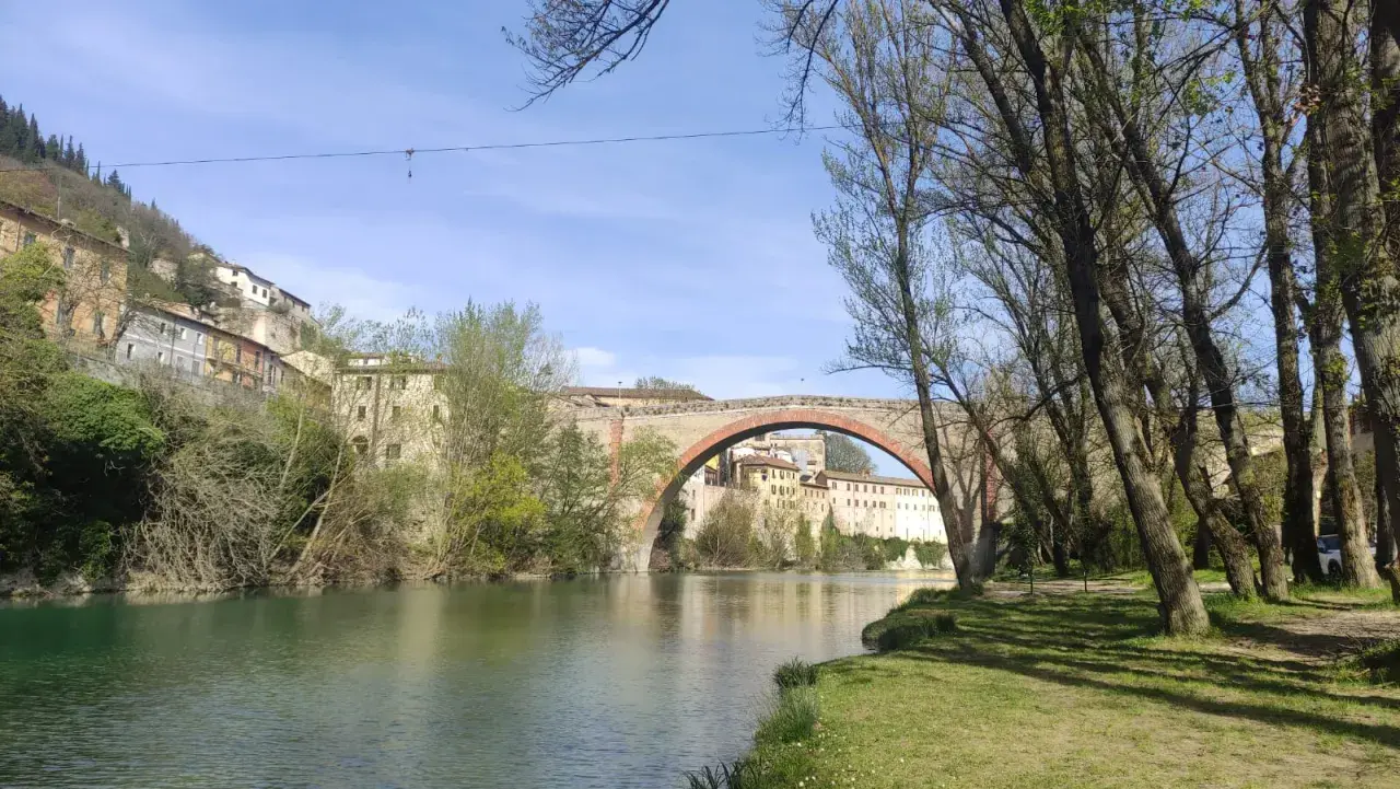 Il fiume scorre placido sotto il ponte ad arco del canyon di Fossombrone, con case antiche aggrappate alla collina e alberi che incorniciano la scena.