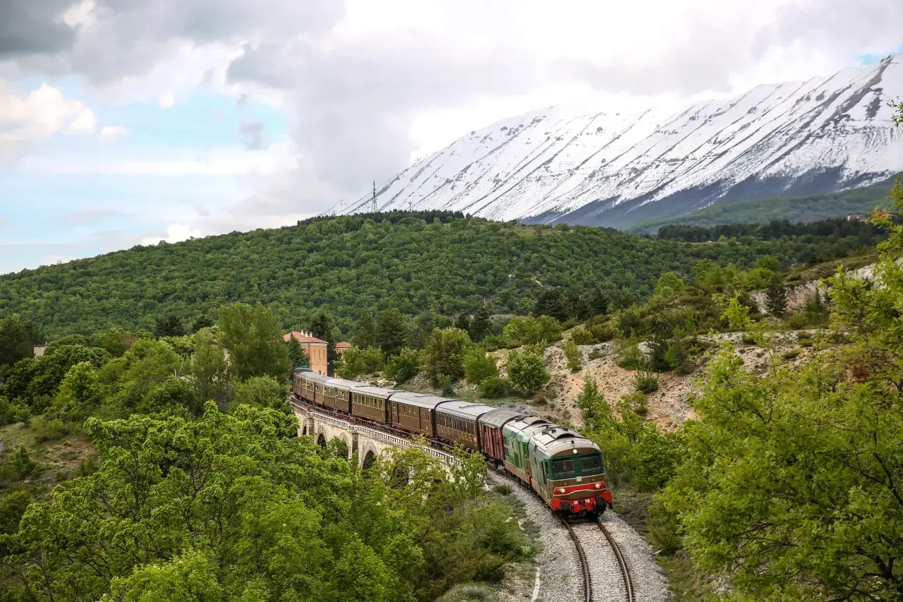 Treno storico attraversa un ponte in pietra, circondato da verde lussureggiante e montagne innevate. Un viaggio suggestivo sulla Transiberiana d'Italia.
