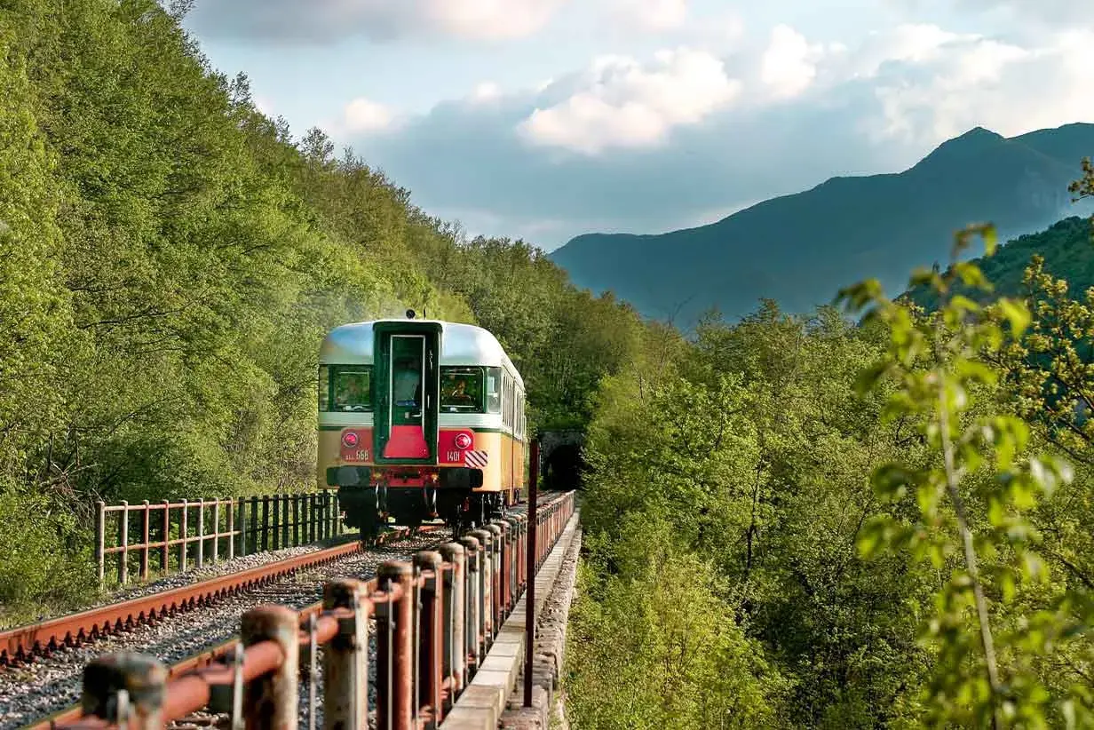 Treno d'epoca attraversa un viadotto immerso nel verde, tra Fabriano e Ancona, dirigendosi verso un tunnel tra le montagne.