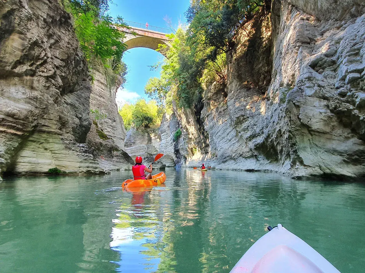 Persone in canoa navigano un fiume tra alte pareti rocciose, un paesaggio che ricorda le marmitte dei giganti, sotto un antico ponte ad arco.