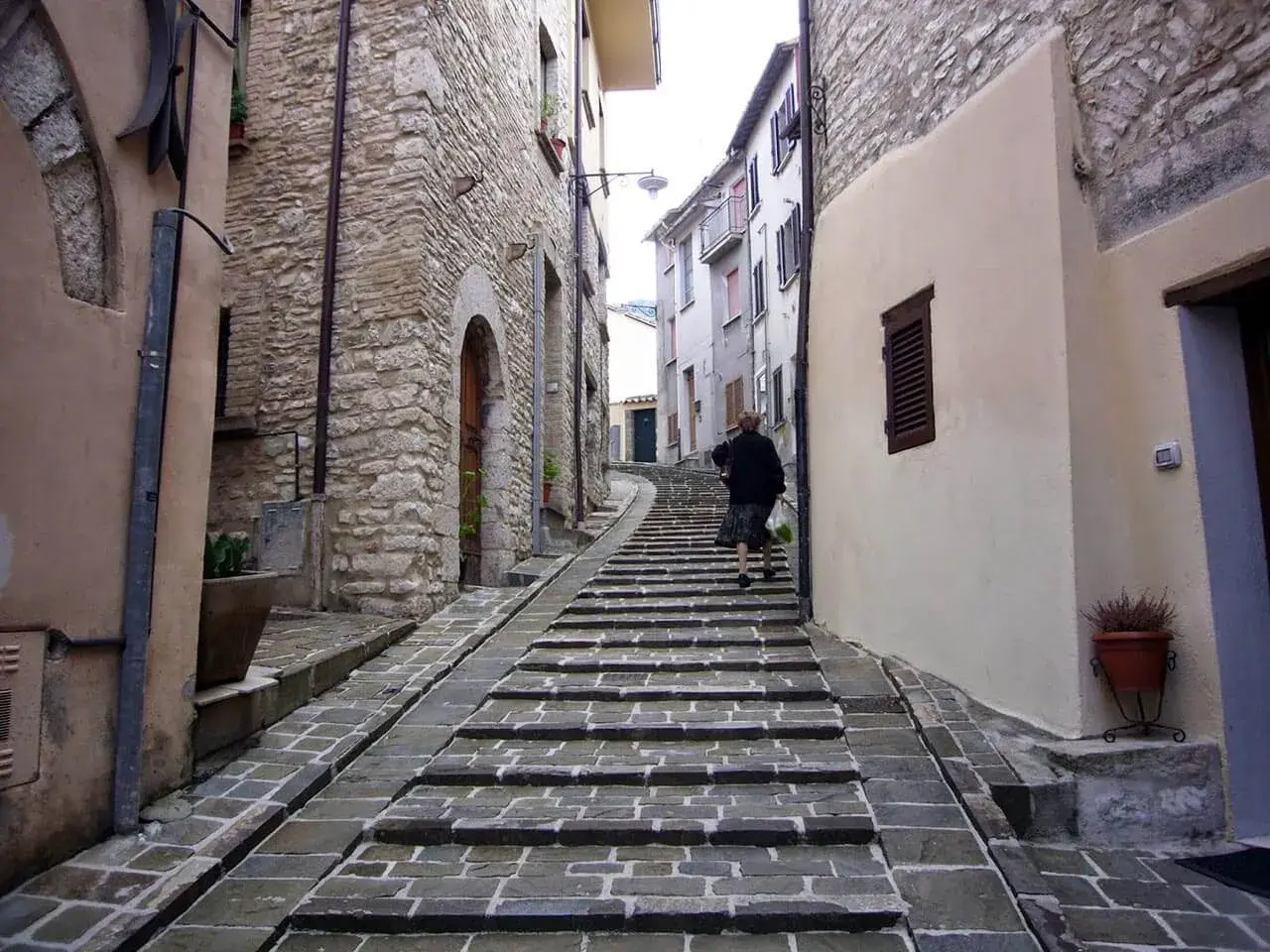 Una donna sale una scalinata in pietra in un vicolo di Gualdo Tadino, tra antiche case in pietra e intonaco.